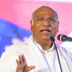 Kharge wearing glasses and a white shirt speaks passionately at a microphone, addressing caste violence in India. He gestures with one hand, highlighting the urgent need for change against a blurred, colorful background.