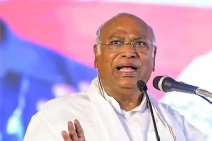 Kharge wearing glasses and a white shirt speaks passionately at a microphone, addressing caste violence in India. He gestures with one hand, highlighting the urgent need for change against a blurred, colorful background.