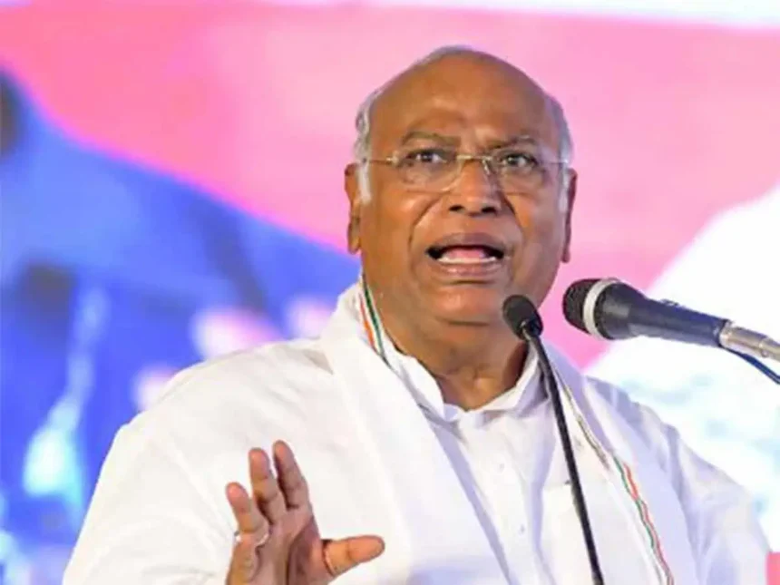 Kharge wearing glasses and a white shirt speaks passionately at a microphone, addressing caste violence in India. He gestures with one hand, highlighting the urgent need for change against a blurred, colorful background.