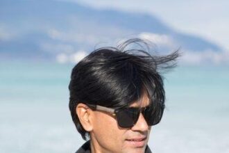 Mohammed Zubair wearing sunglasses and a black jacket stands in front of a blue sea, reminiscent of the independent journalism in India, where clarity cuts through blurred mountains of misinformation. The wind tousles their hair as they look slightly to the side.