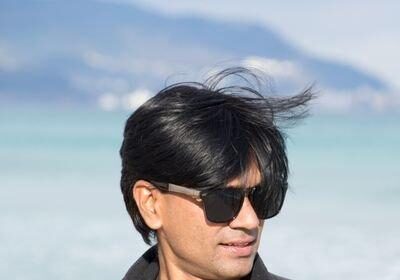 Mohammed Zubair wearing sunglasses and a black jacket stands in front of a blue sea, reminiscent of the independent journalism in India, where clarity cuts through blurred mountains of misinformation. The wind tousles their hair as they look slightly to the side.